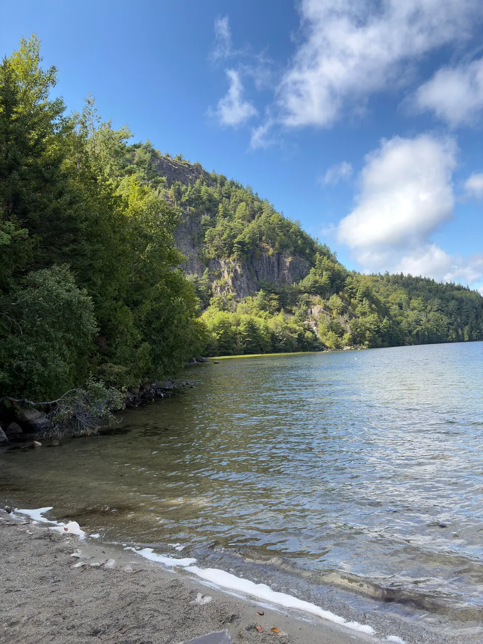 A small sandy beach with clear rippling water looks along a lake to a steep wooded granite cliff under a blue sky with white clouds.