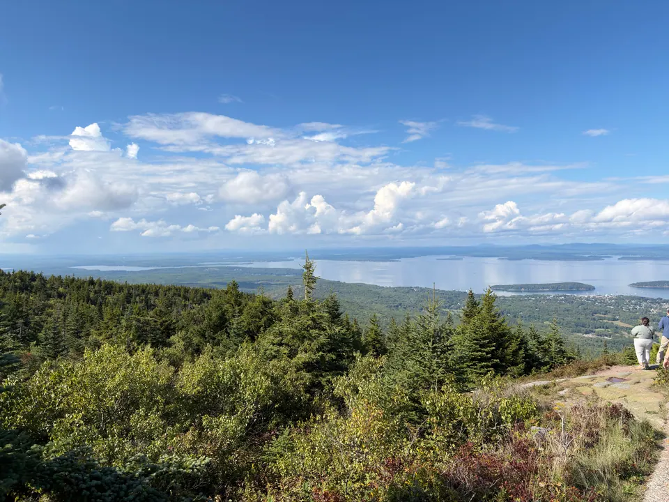 A wide view from a mountaintop over green spruce scrub down to Frenchman Bay, with islands in the water and puffy white clouds in a blue sky; two hikers walk a granite path at the right.