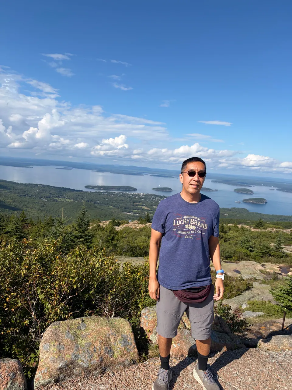 A man in sunglasses and a blue T-shirt stands on pink granite at a summit, with a bay full of small wooded islands spread out below him.