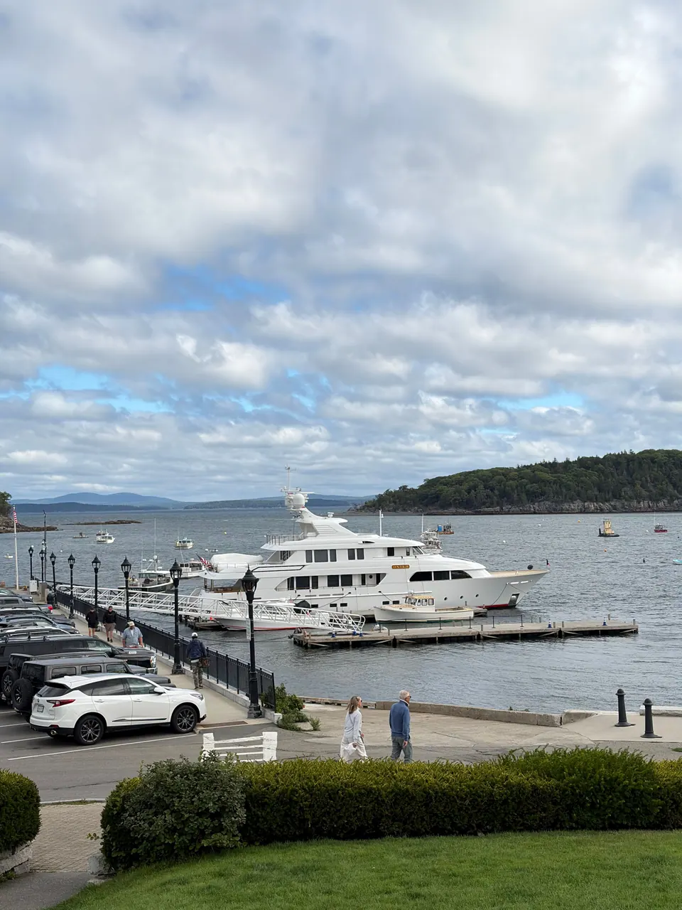 Looking over a lawn and a parking lot to a pier where a large white motor yacht is moored, with a wooded island across grey water under a mottled cloudy sky.