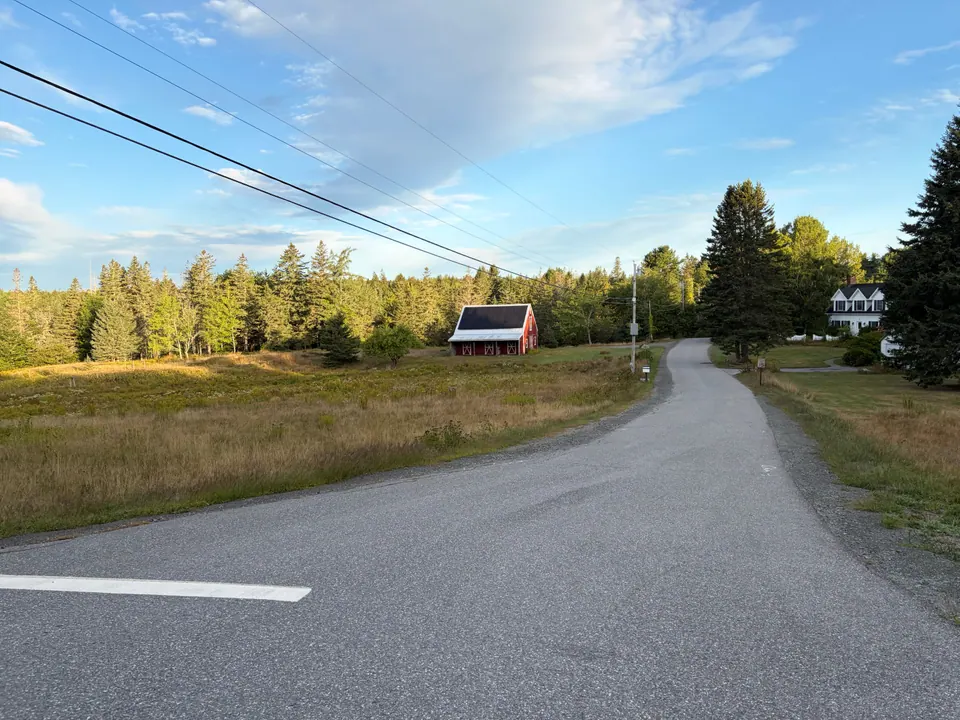 Early-morning light on a quiet country road curving past a hay meadow, a small red barn and a white farmhouse, with spruce trees and power lines under a pale blue sky.