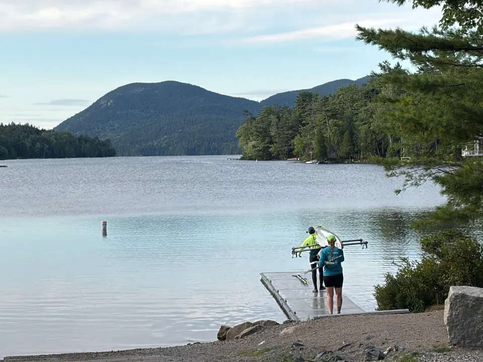 A still lake with a rounded wooded mountain across the water; two people in bright jackets carry a narrow boat along a small floating dock.