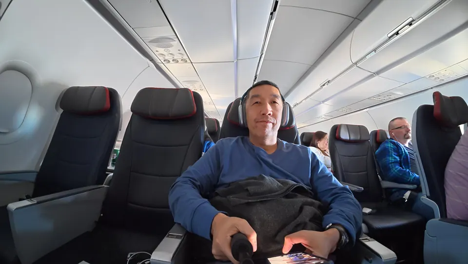 A smiling man in a blue sweater and headphones sits in a dark leather airplane seat, a jacket on his lap, other passengers behind him.