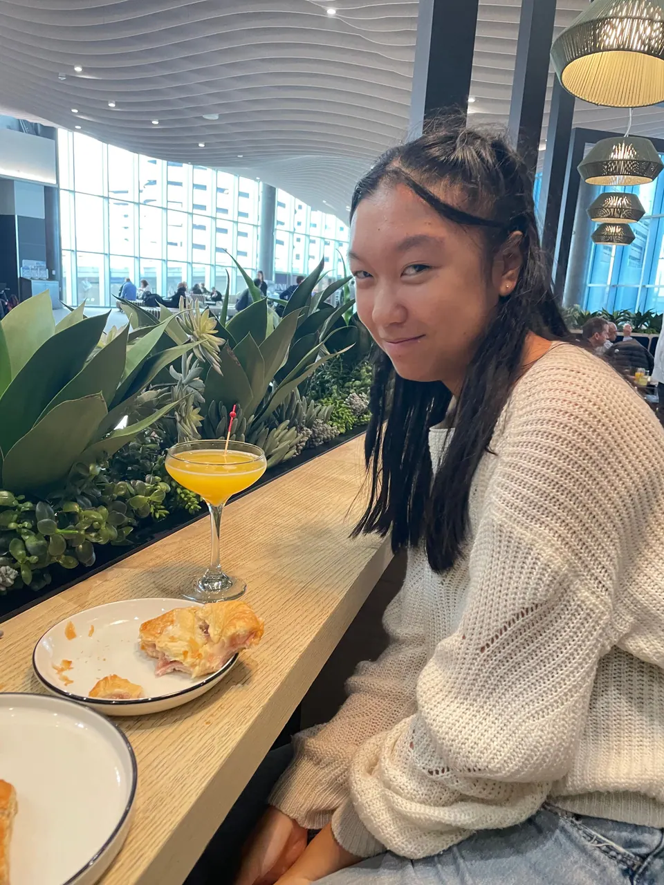 A girl in a cream knit sweater sits at a long wooden counter lined with agave plants, a glass of orange juice and a half-eaten pastry in front of her, big windows behind.