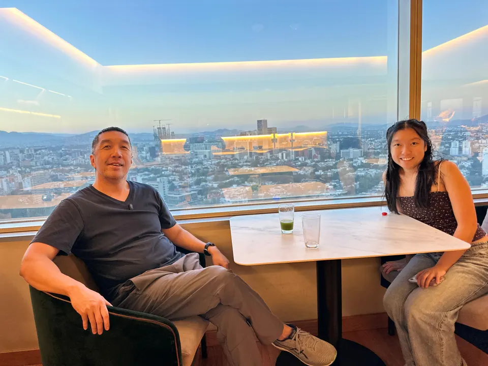 A man and a girl sit at a small marble table by a wide window high above a city that spreads to the hills under a pale evening sky.