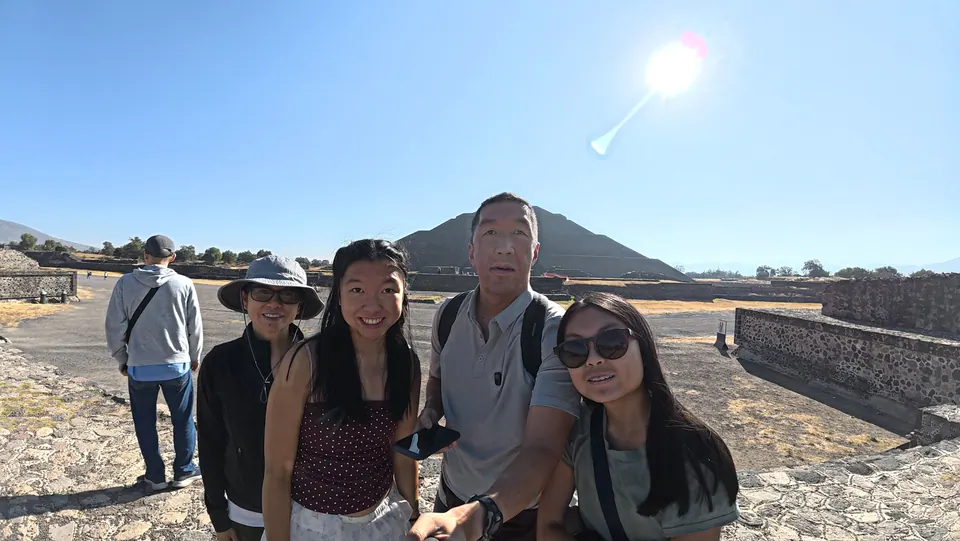 Four people smile in a selfie on the stone-paved Avenue of the Dead under a deep blue sky, the sun flaring above; a great stepped pyramid rises behind them and low stone platforms line the avenue.