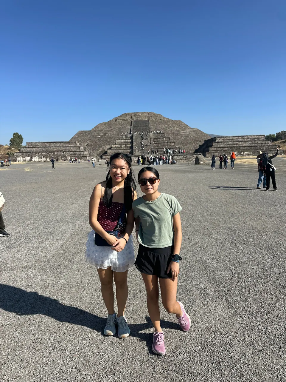 Two girls stand arm in arm on a wide gravel avenue under a cloudless sky, with a large stepped stone pyramid and a crowd of visitors behind them.