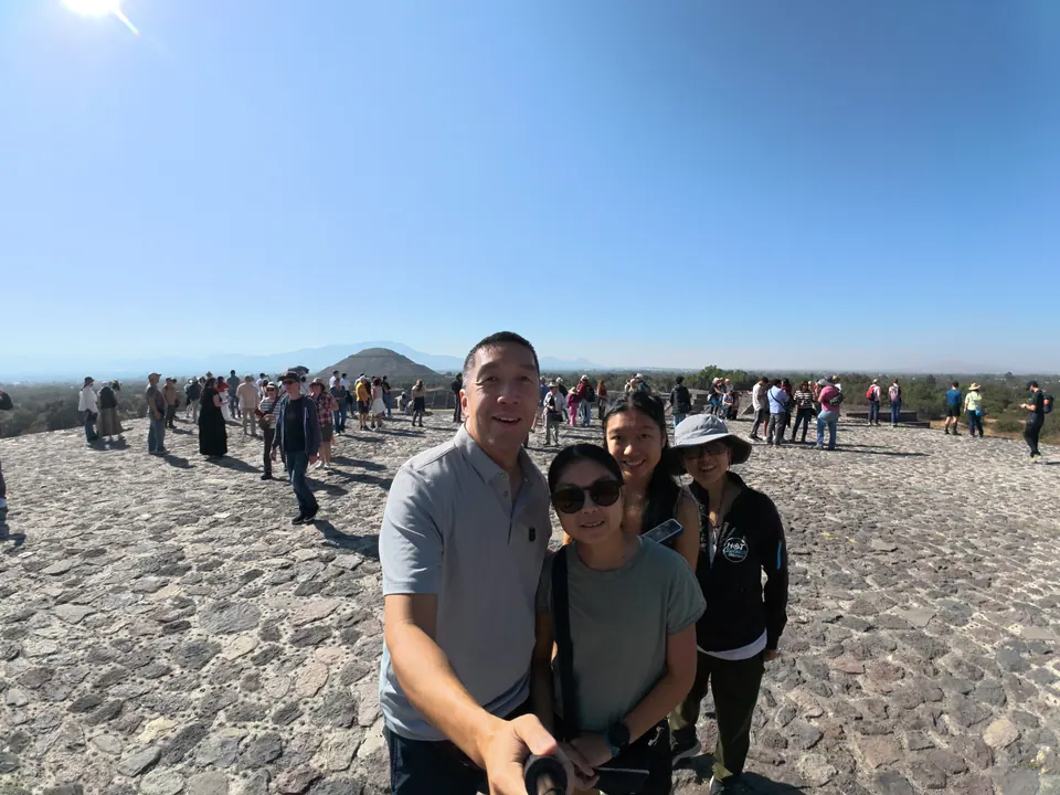 A man holding a selfie stick and three women and girls smile on a wide stone summit crowded with visitors, a flat valley, a smaller pyramid and hazy mountains far behind under a blue sky.