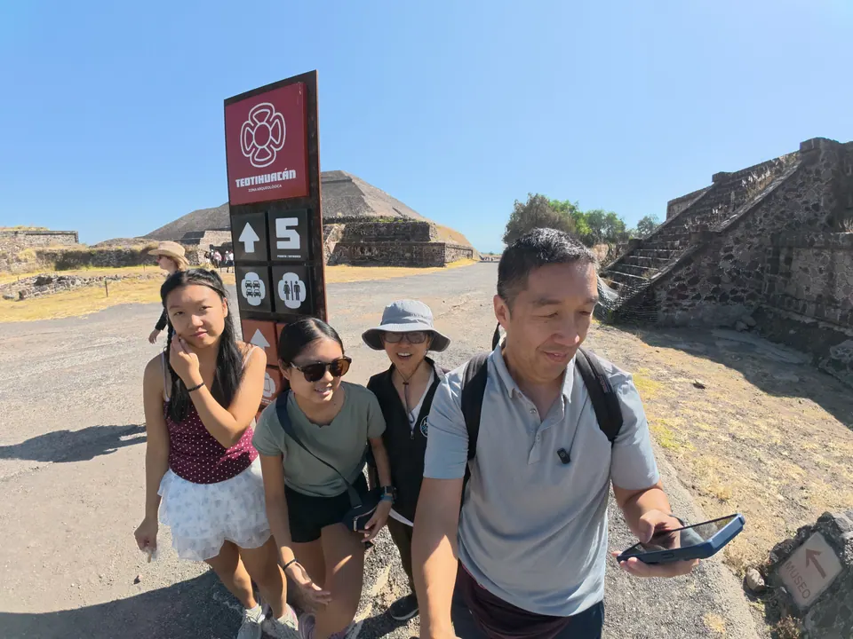 Four people walk past a tall red and dark sign reading Teotihuacán with an arrow and the number 5; a large stepped pyramid is behind them and stone steps rise at the right.