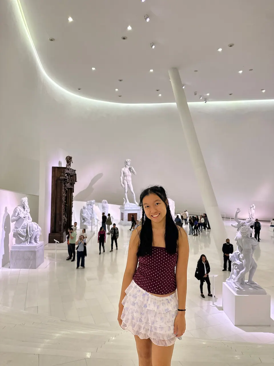 A smiling girl stands in a bright white gallery with a curving ceiling and white marble statues, including a copy of Michelangelo's David, behind her.