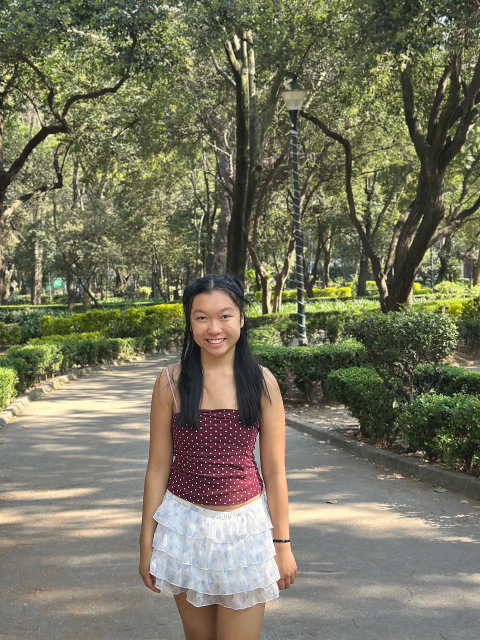 A girl in a polka-dot top and a white ruffled skirt stands on a shady park path between clipped hedges and tall trees.
