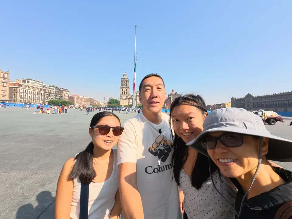 Four smiling people take a selfie on a vast paved square under a clear sky; a tall flagpole with a Mexican flag, a cathedral bell tower and colonial buildings are behind them.
