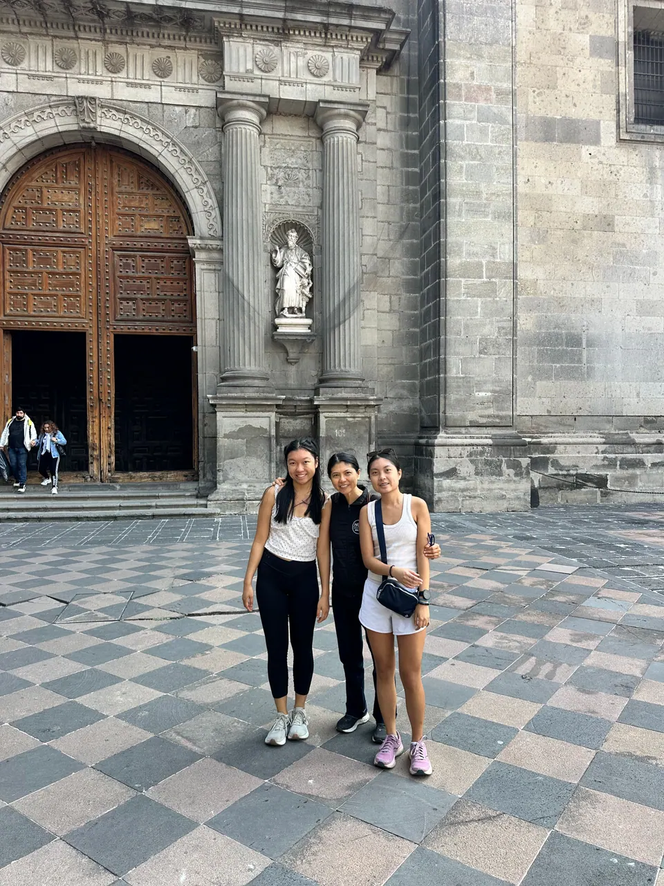 Three people stand on black-and-white checkered paving in front of a carved stone doorway with tall wooden doors and a statue of a saint in a niche.
