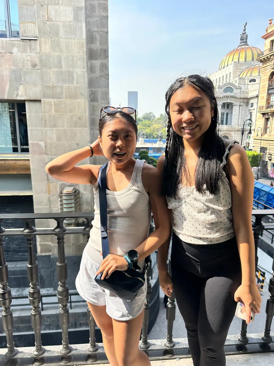 Two girls pose on a balcony with an iron railing in bright sun; a building with a golden-tiled dome rises behind them.