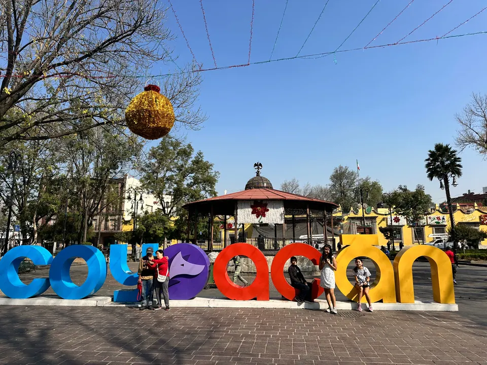 Large brightly colored letters spelling Coyoacán stand in a sunny plaza with people posing among them; a bandstand with a red roof, palm trees and a hanging gold ornament are behind.