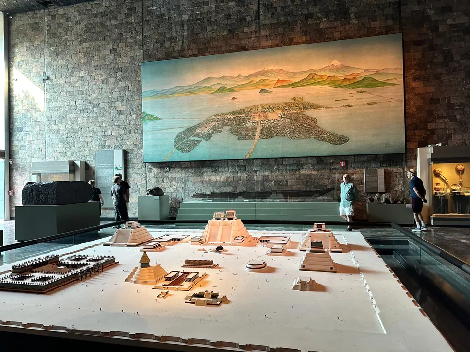 A large tabletop model of a walled temple precinct with pyramids, and behind it a wall-sized painting of an island city in a lake ringed by mountains.