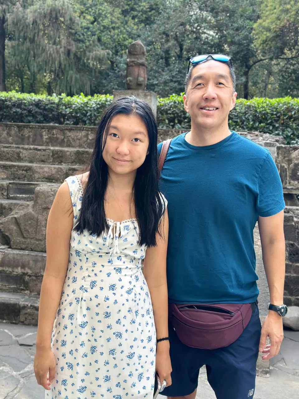 A man in a blue T-shirt and a girl in a flowered dress stand in front of old stone steps and hedges, a carved stone figure behind them.