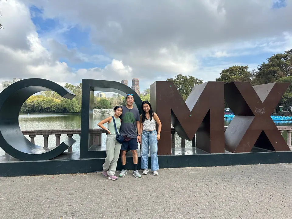 A man and two girls stand inside giant dark-bronze letters spelling CDMX beside a lake with a stone balustrade, trees and a few tall buildings behind, under a cloudy sky.