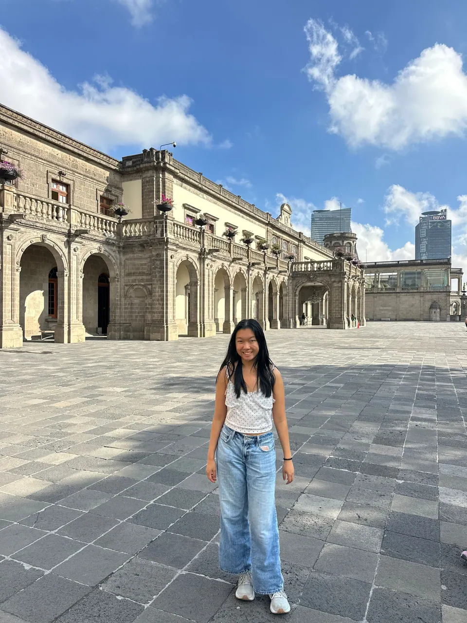 A girl in jeans stands on a large stone-paved courtyard in front of a pale stone palace with arched arcades and balconies, two modern towers beyond under a blue sky with white clouds.