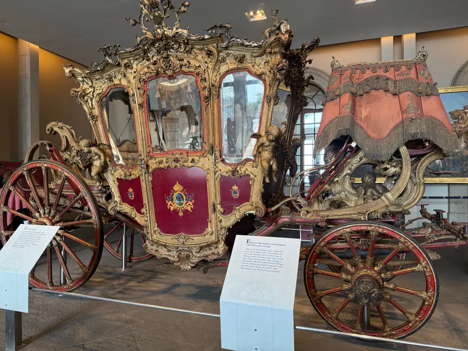 An ornate gilded carriage with red panels, glass windows, carved cherubs and large red spoked wheels on display in a museum hall, with information placards in front.