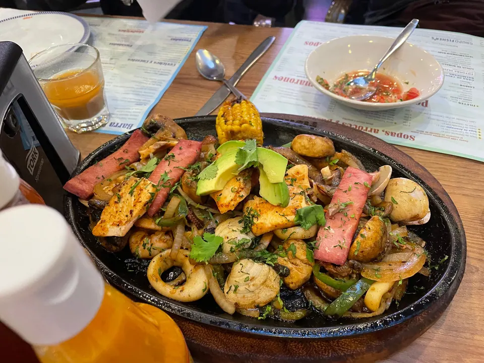 A sizzling black platter of seafood, sliced meat, grilled chicken, mushrooms, peppers, corn and avocado on a restaurant table, with a bowl of salsa and orange drinks around it.