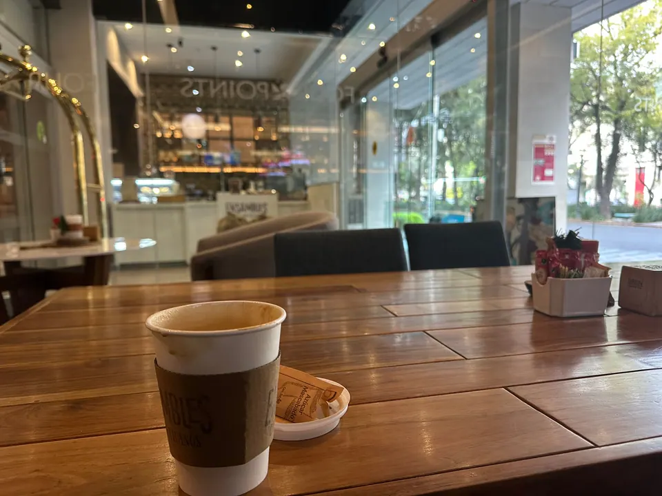A paper cup of coffee in a brown sleeve and a small packet on a saucer sit on a wooden table in a quiet café, with glass walls looking out on a tree-lined street.
