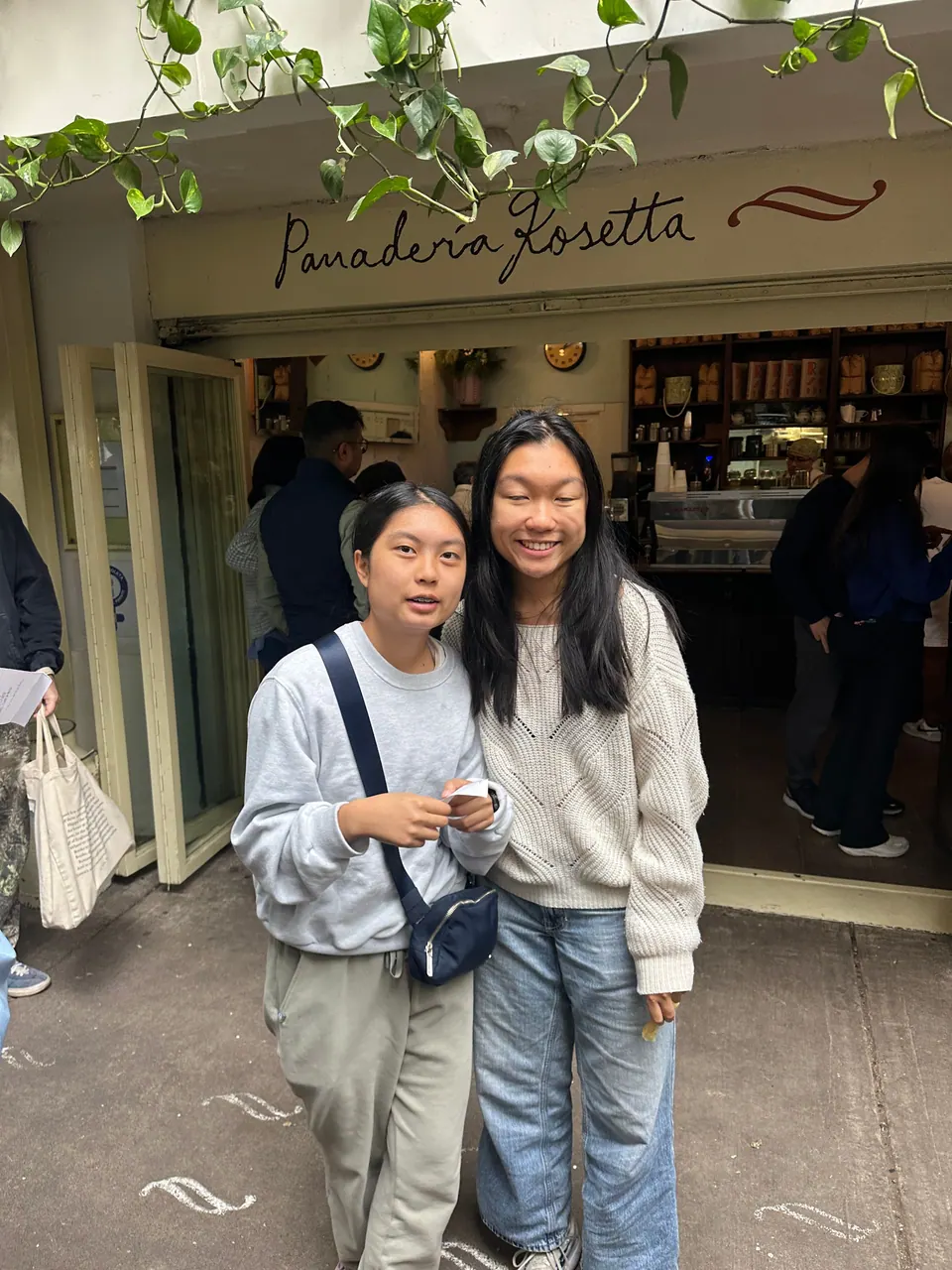 Two girls in sweaters stand close together in front of a bakery doorway under a hand-lettered sign reading Panadería Rosetta, with vines above and customers inside.
