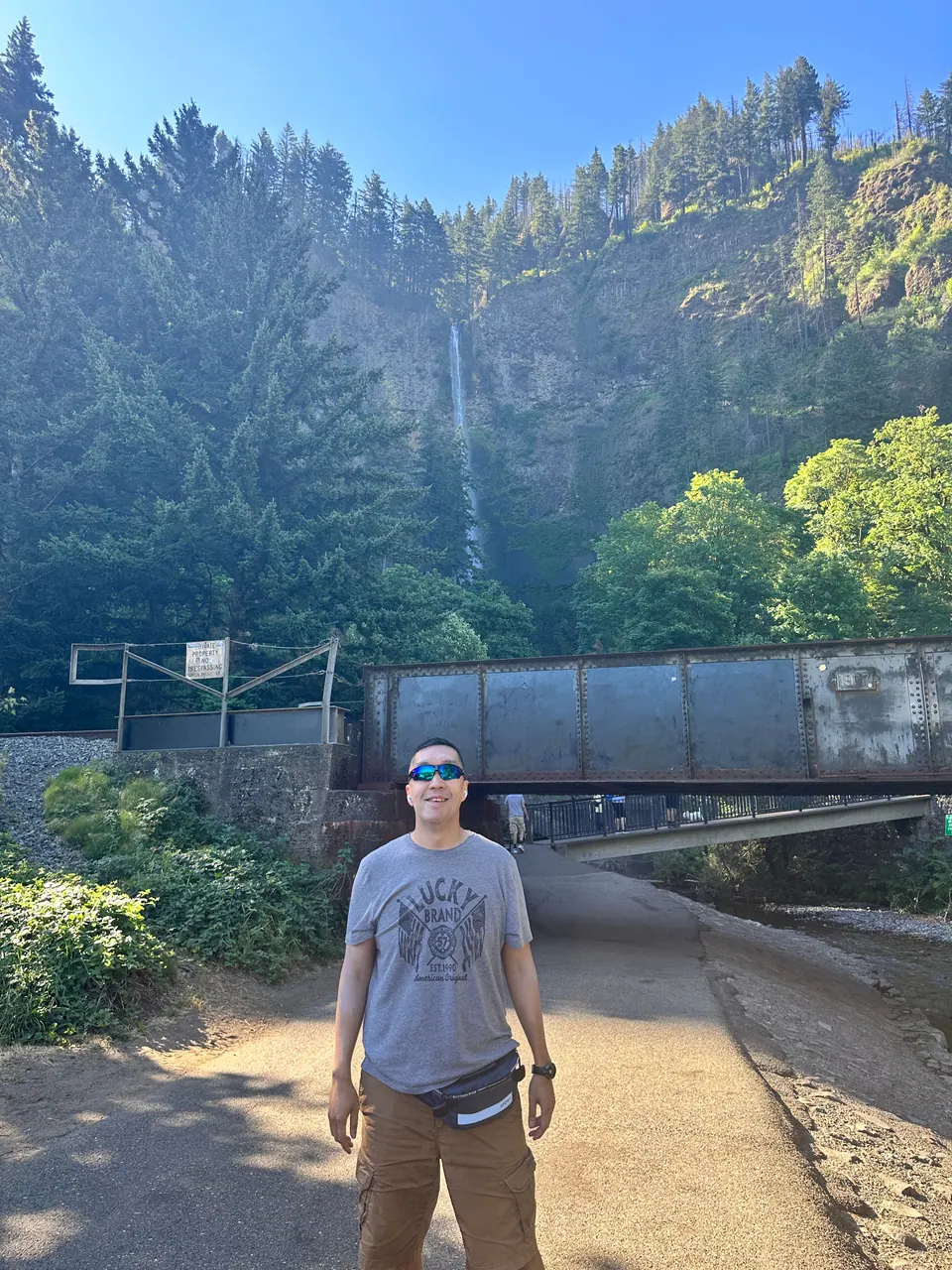 A man in a gray T-shirt and sunglasses stands on a sunny path; a tall, thin waterfall and a stone bridge are behind him.
