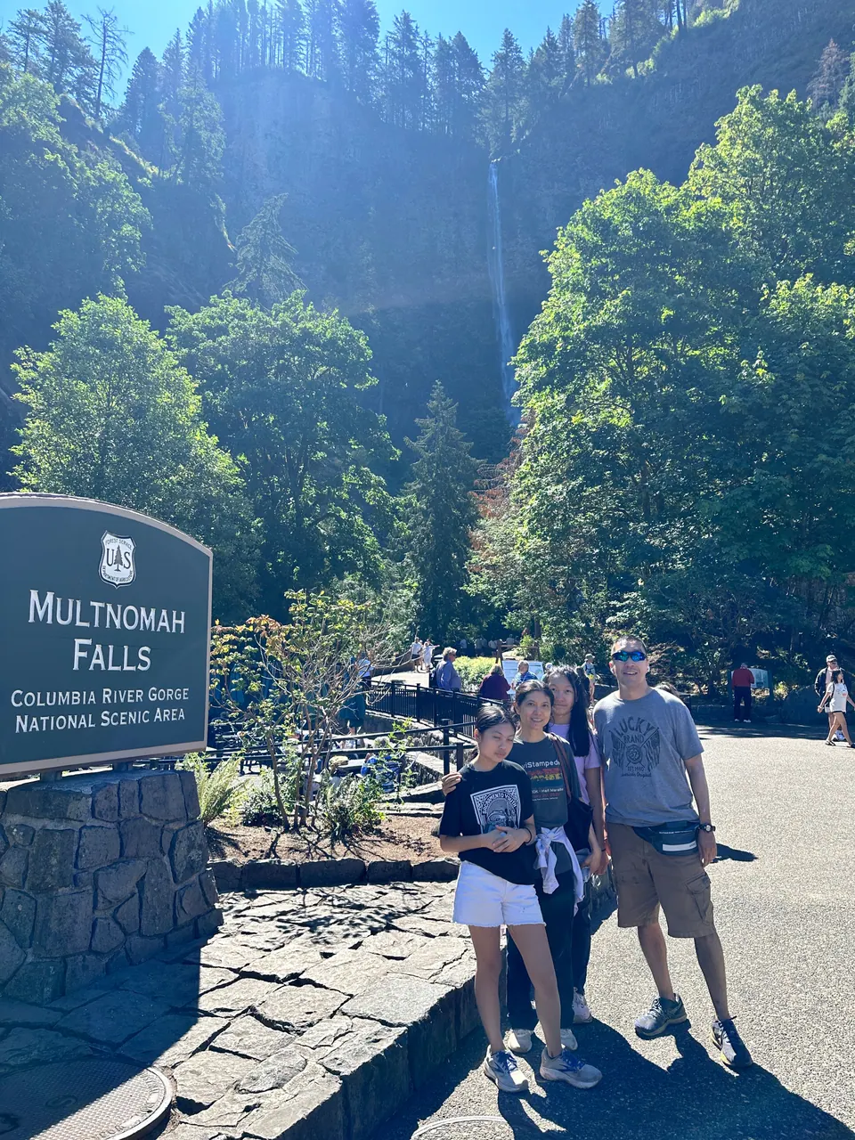 Three people pose beside a stone sign reading Multnomah Falls, with forested cliffs behind them.