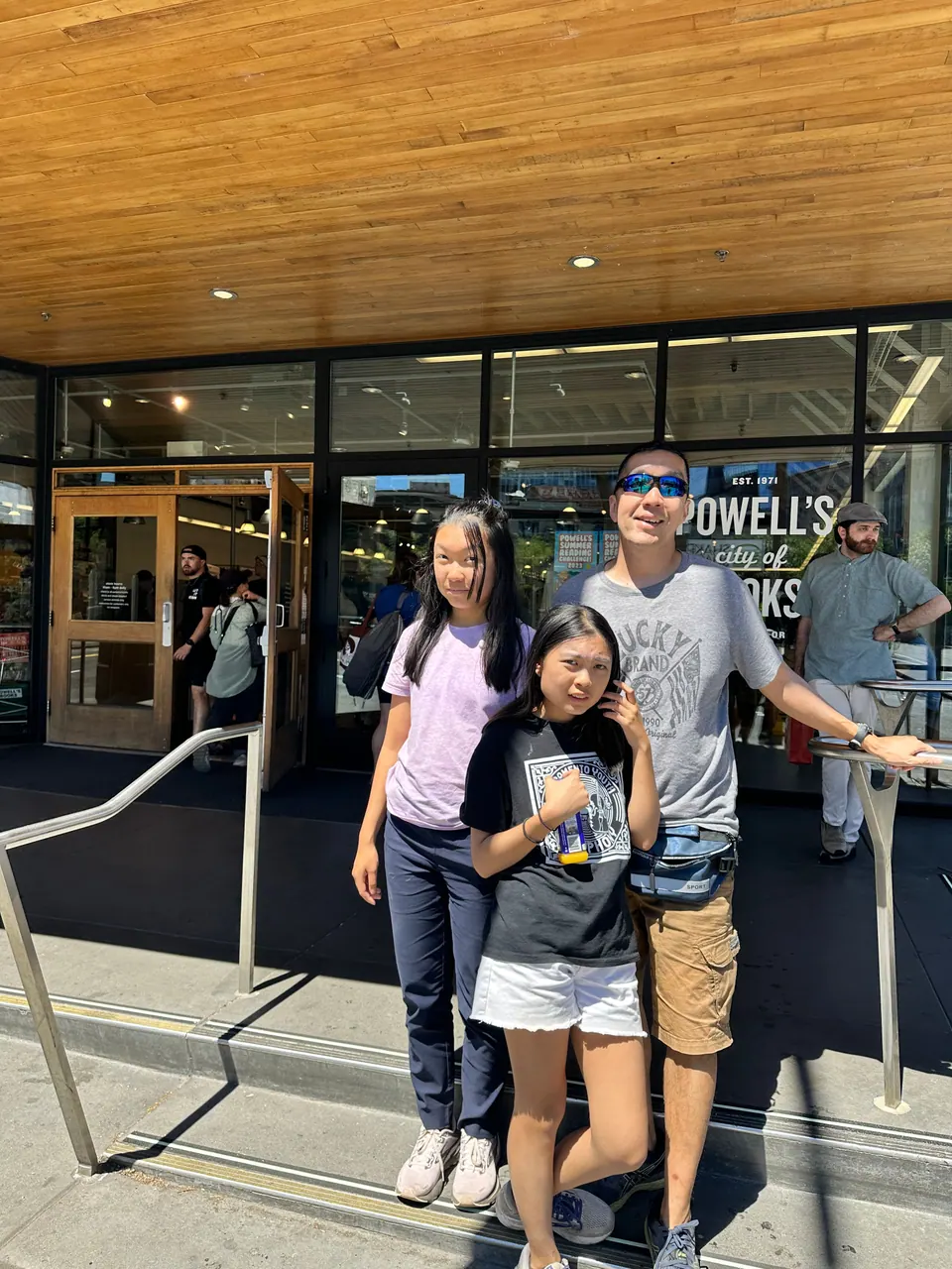 Two girls and a man stand at the entrance of a bookstore under a wood-slatted ceiling; the sign reads Powell's.