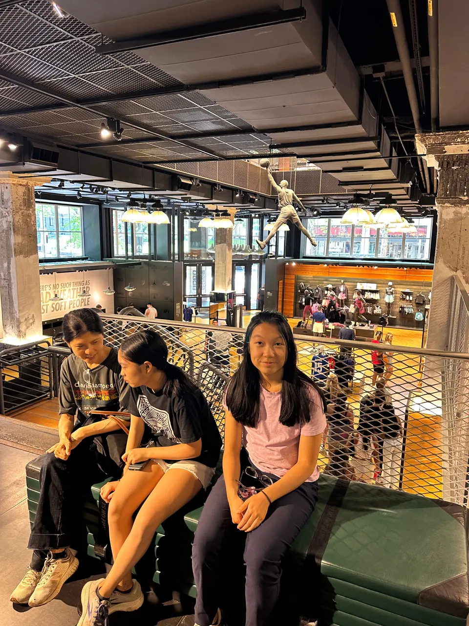 Three people sit side by side on a green bench on an upper floor of a large bookstore, with a railing behind them.