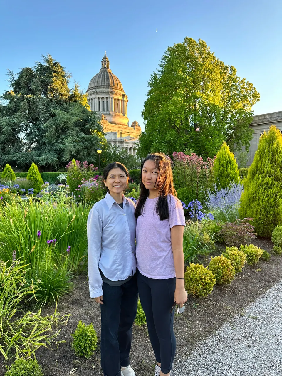 Two people stand among garden plants and flowering shrubs with a capitol dome and trees behind them.