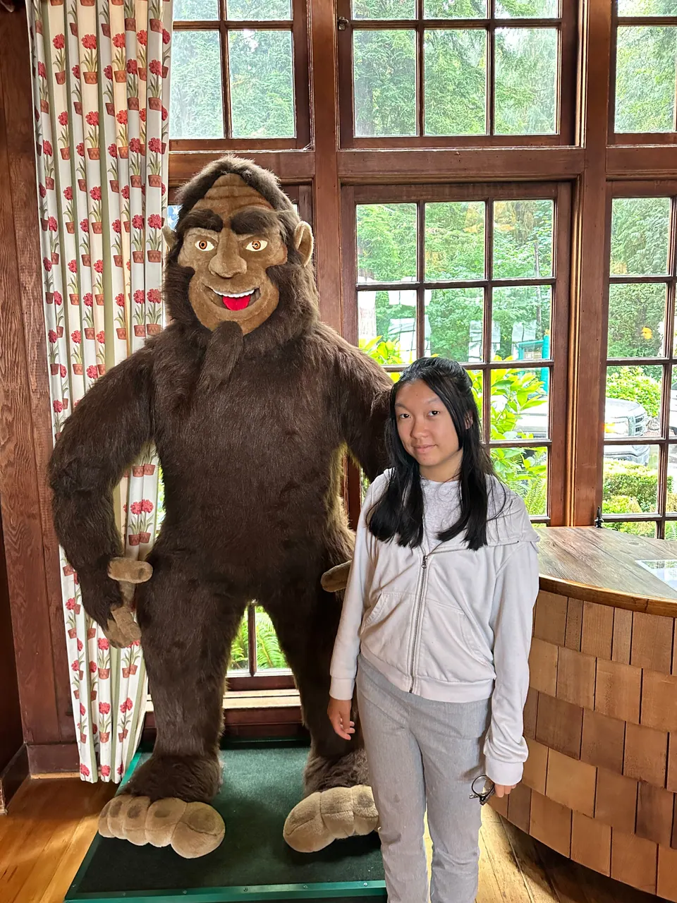 A girl stands next to a tall bigfoot figure in front of a paned window.