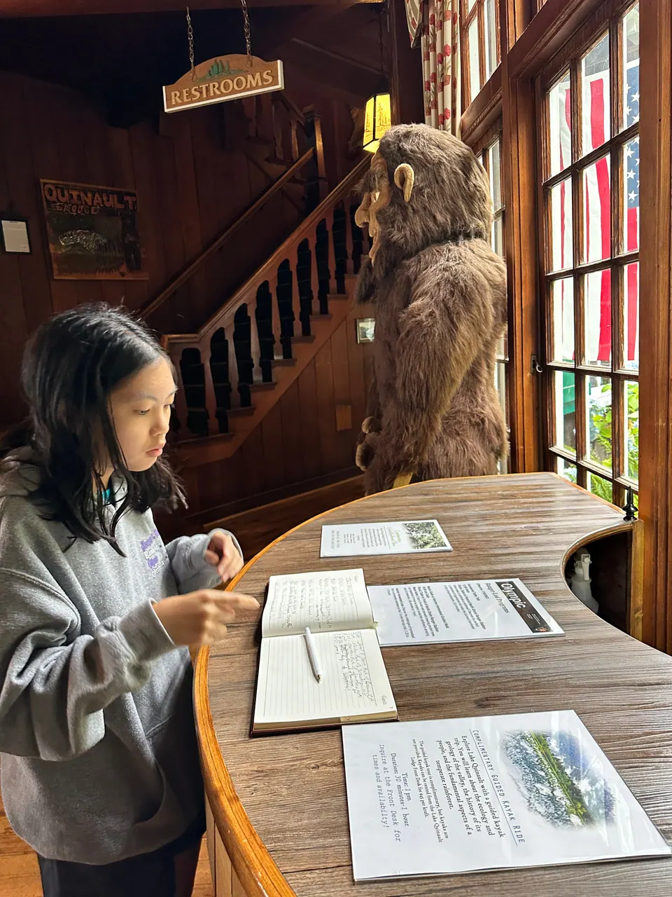 A girl reads papers laid out on a curved wooden counter; a large bigfoot figure stands near a staircase behind her.