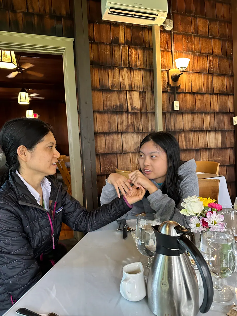 Two people sit at a table with a coffee carafe and a small vase of flowers, a shingled wall behind them.