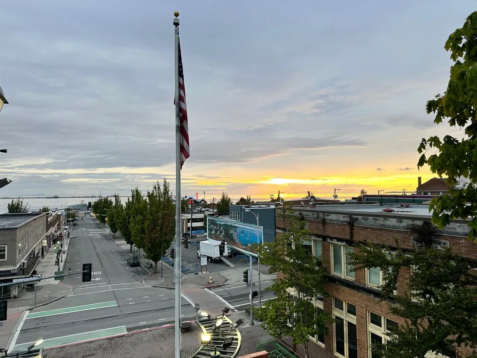 Dawn light over a small town's main street, with a flagpole and low buildings and the sea on the horizon.