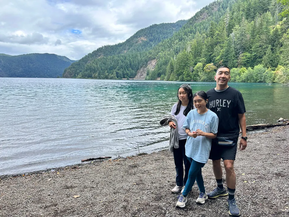 Three people stand on a pebble shore in front of a clear green lake and forested mountains under clouds.