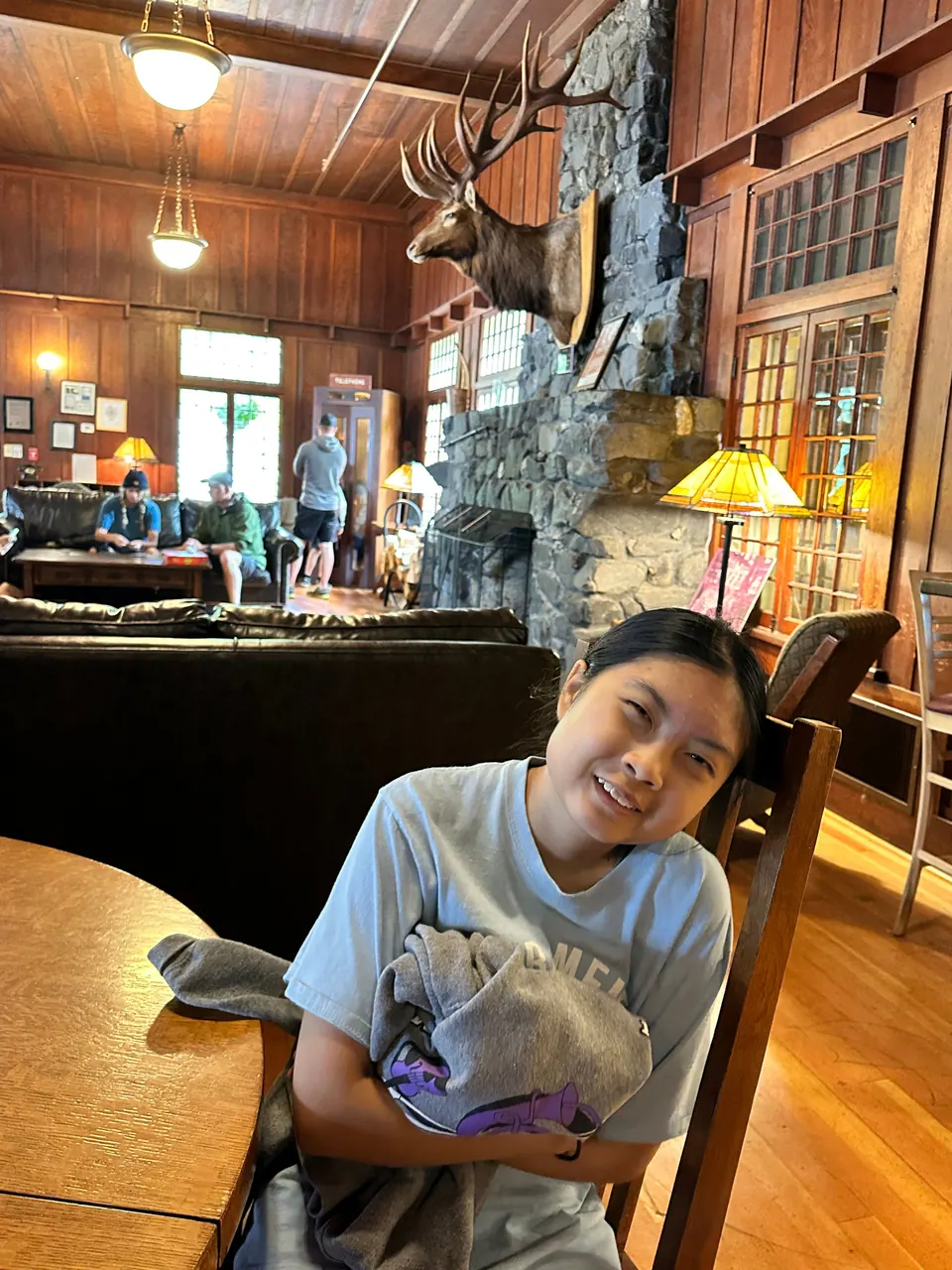 A smiling girl sits in a wooden chair in a lodge lobby with a stone fireplace and mounted antlers.