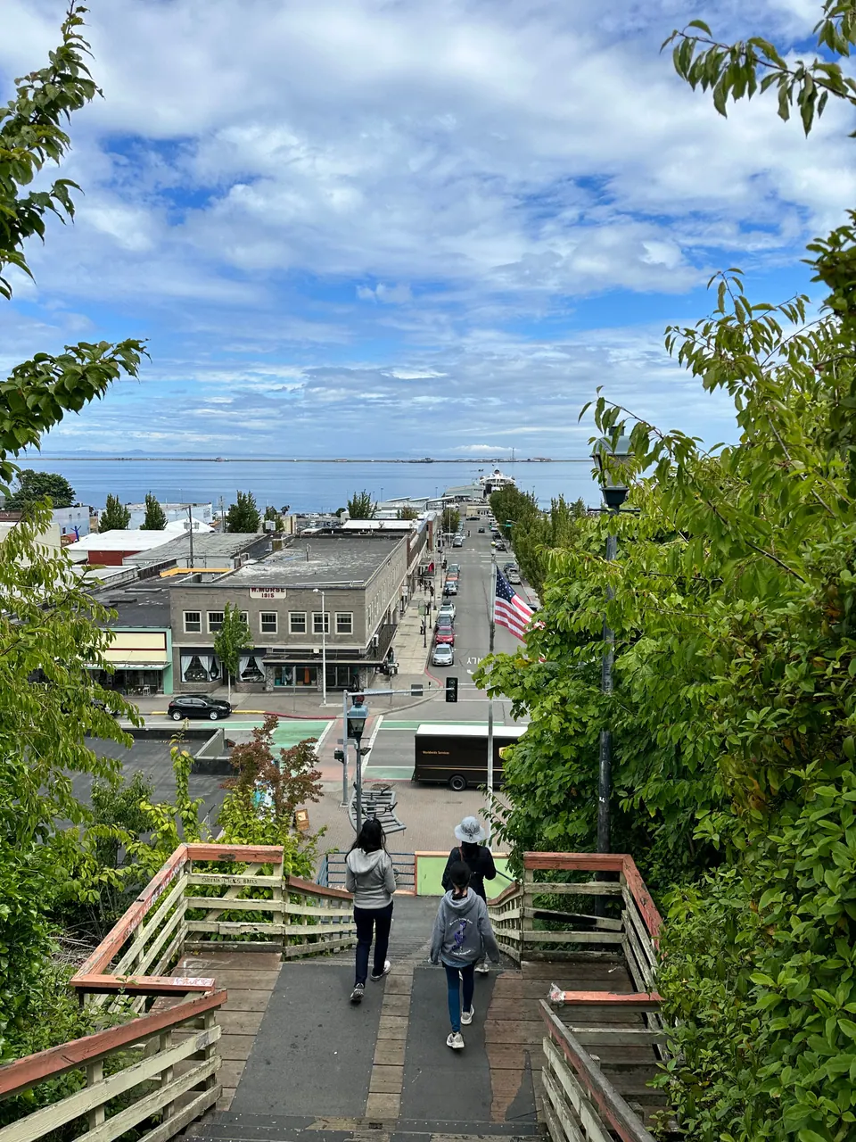 Two people walk down a wooden staircase toward a small town and the sea under a cloudy blue sky.