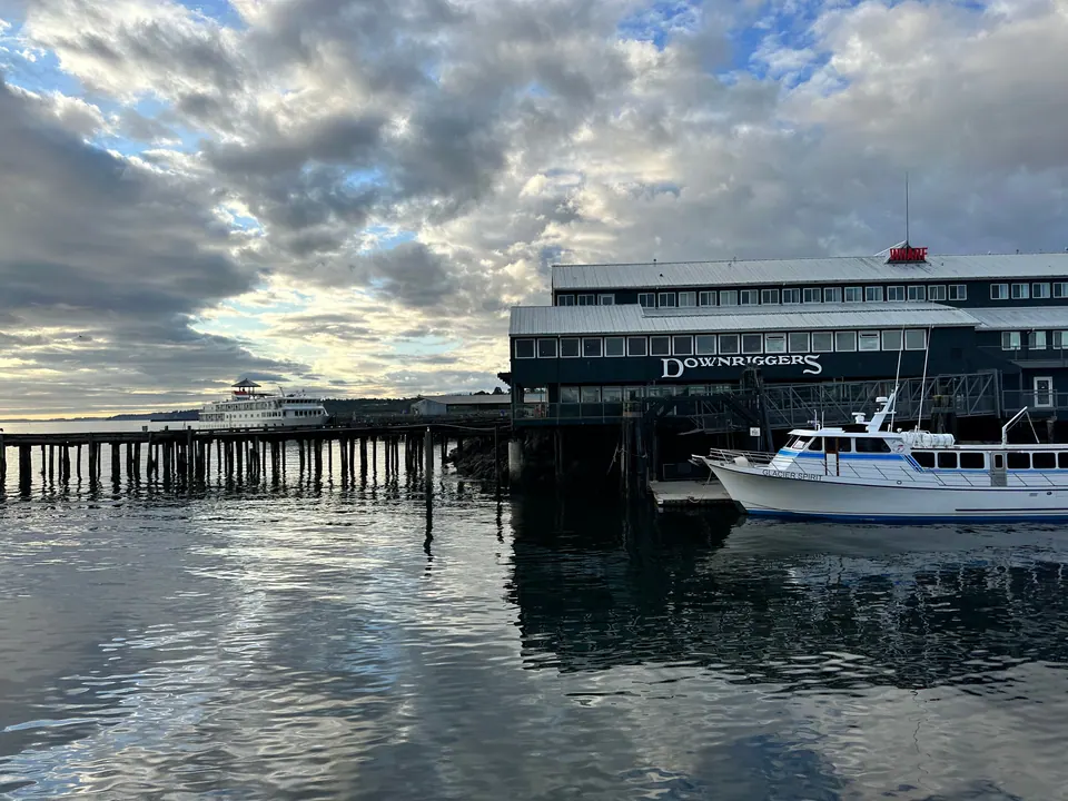 A pier and a waterfront building reflected in calm water under a sky of broken clouds; a white boat is moored alongside.