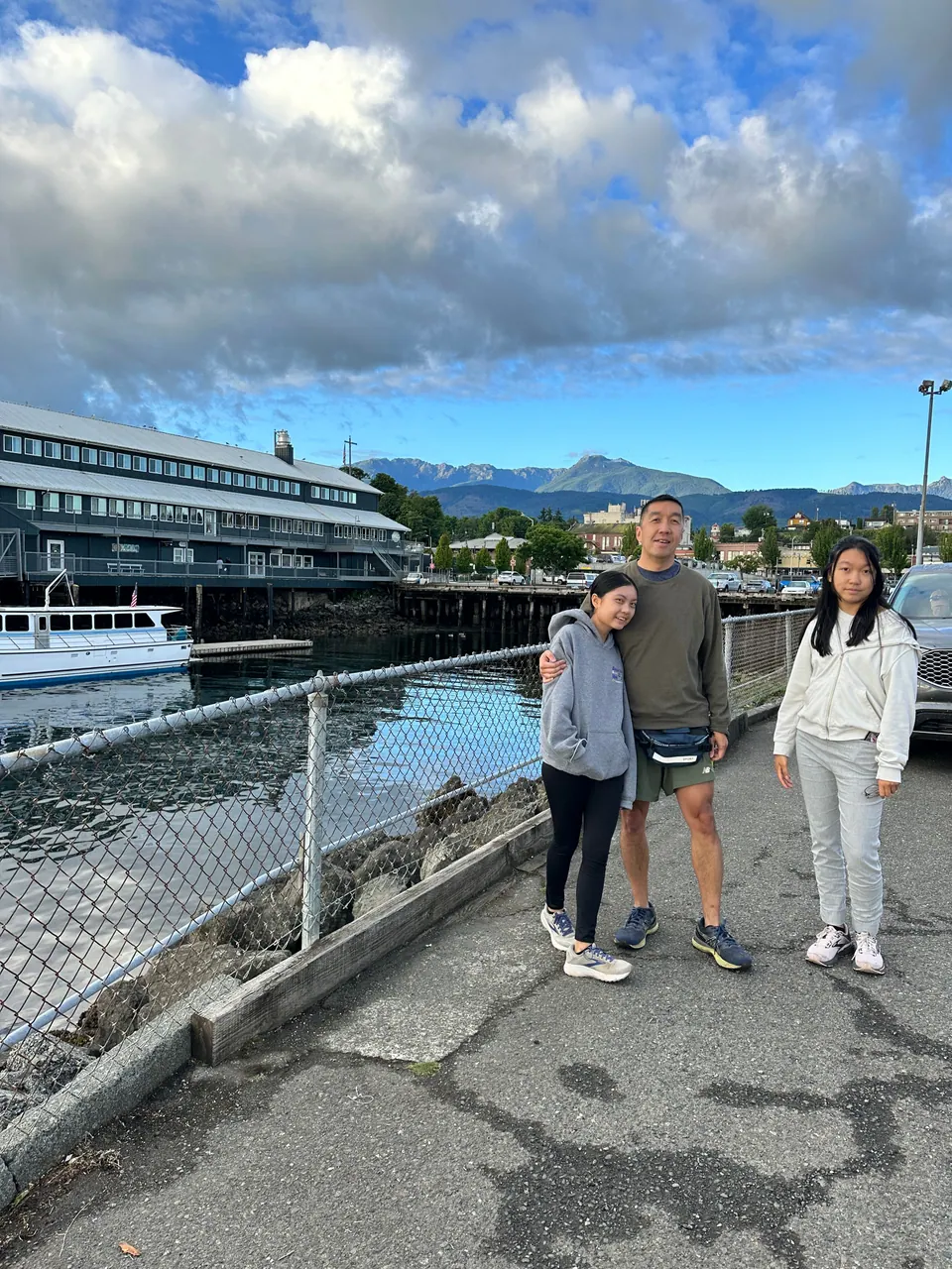 Three people stand by a railing at a harbor, with a terminal building, mountains and clouds behind them.