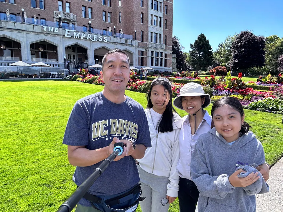 A man holding a selfie stick and three smiling women and girls stand on a lawn in front of a large château-style hotel and flower beds.