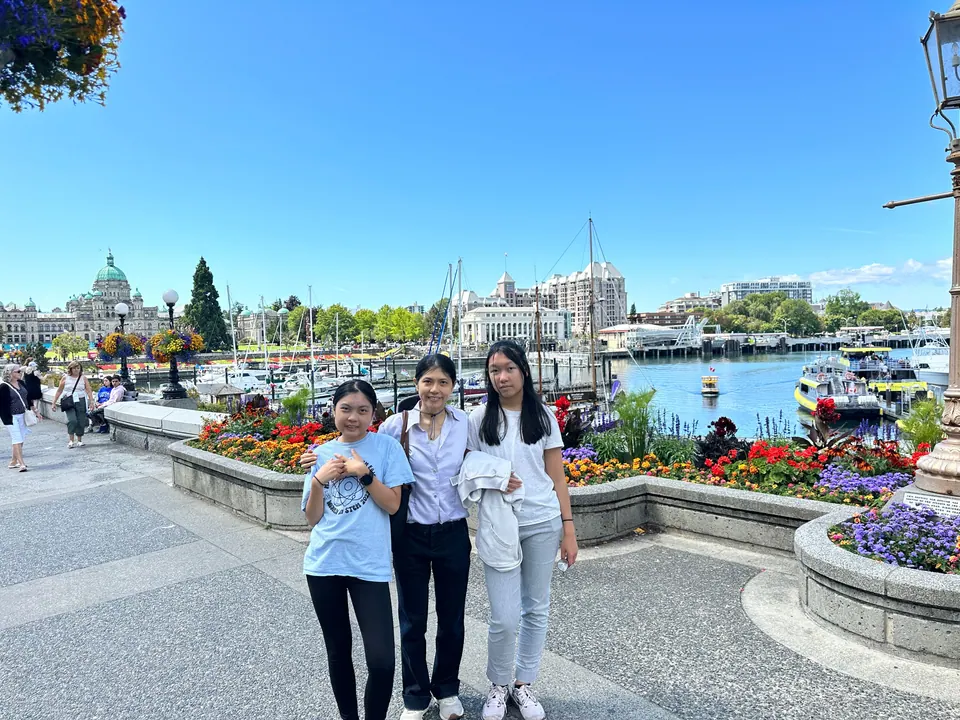 Three people stand by harbor flower beds; a domed legislative building and moored boats are behind them.