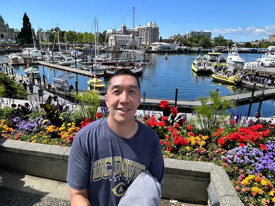 A smiling man in a T-shirt stands in front of red flowers and a marina.