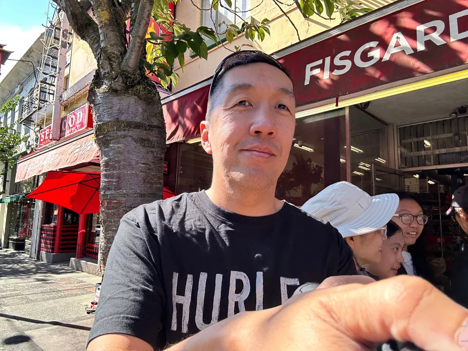A man takes a selfie on a city sidewalk; a red storefront sign reading Fisgard is above him and two people are behind him.