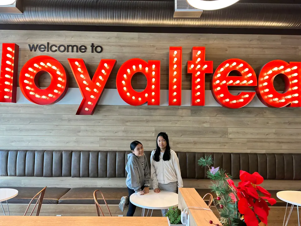 Two girls stand under a large red illuminated sign reading welcome to loyaltea inside a café.