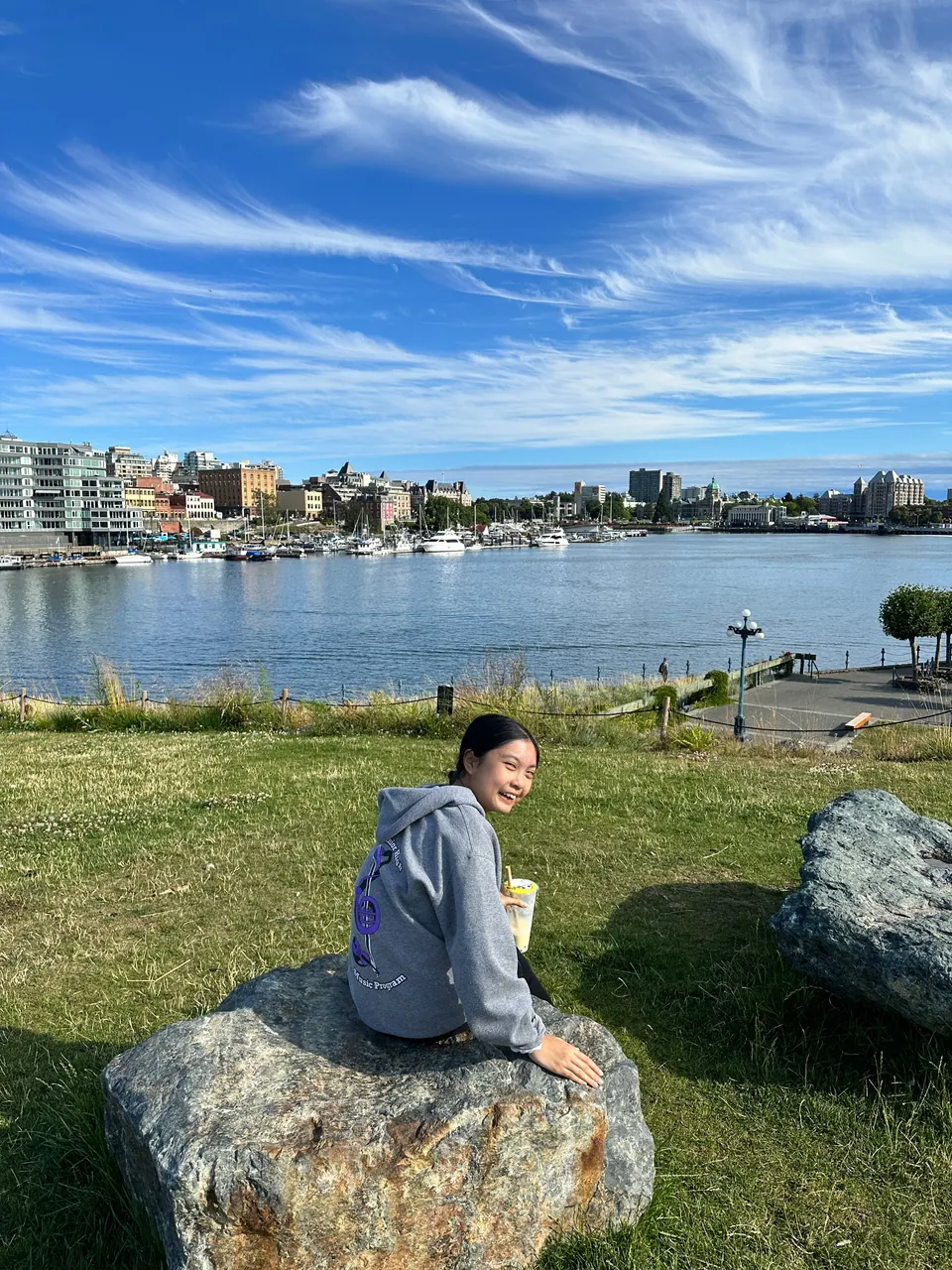 A girl sits on a boulder on a grassy bank above blue water, with buildings across the harbor.