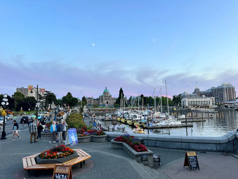 A harbor promenade at dusk with flower beds and people walking, and a domed building across the water.