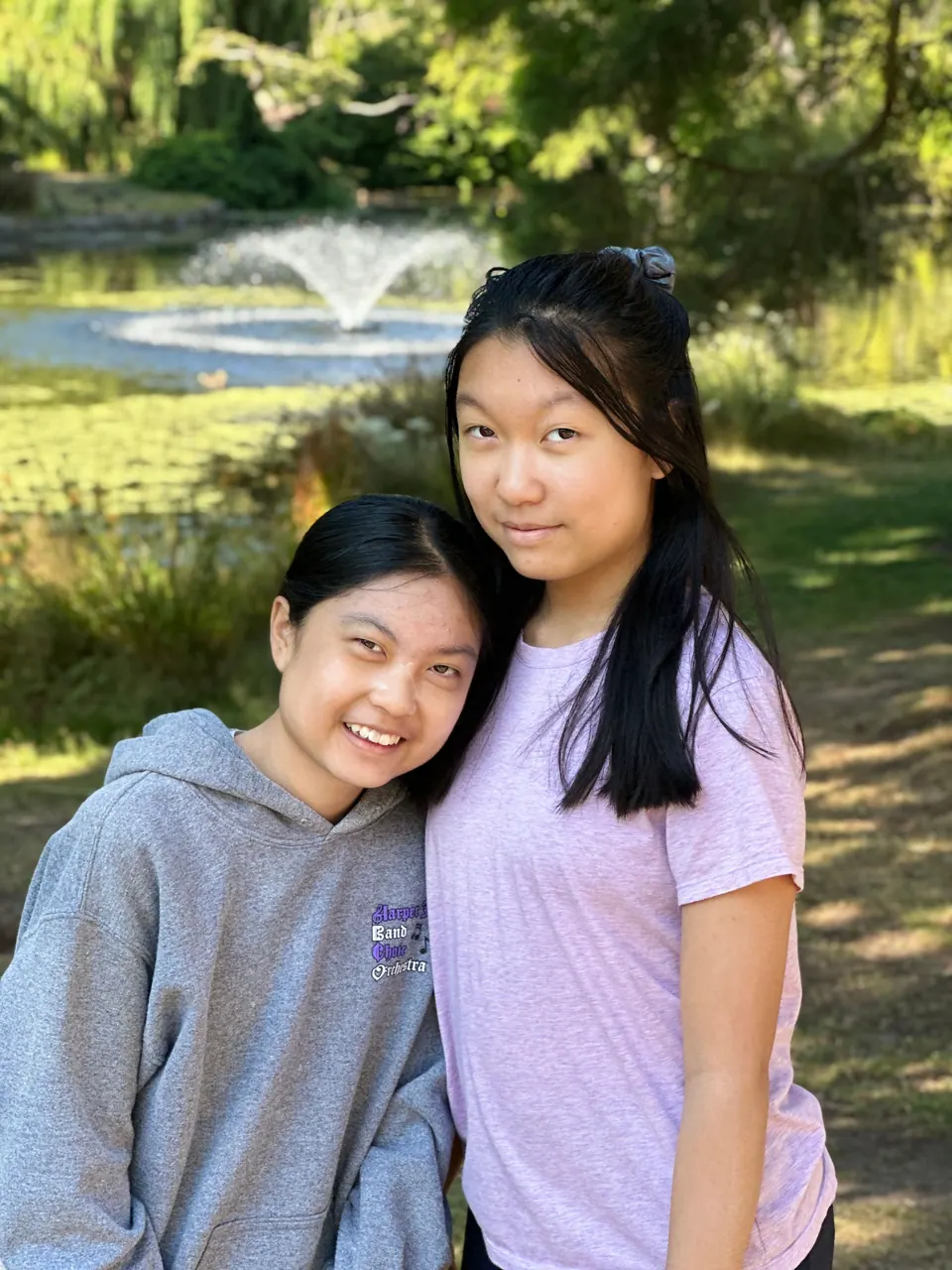 Two smiling girls stand close together, with a pond and a fountain behind them.
