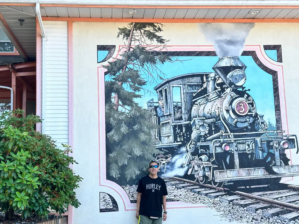 A man in a black T-shirt stands in front of a large painted mural of a steam locomotive on a building wall.